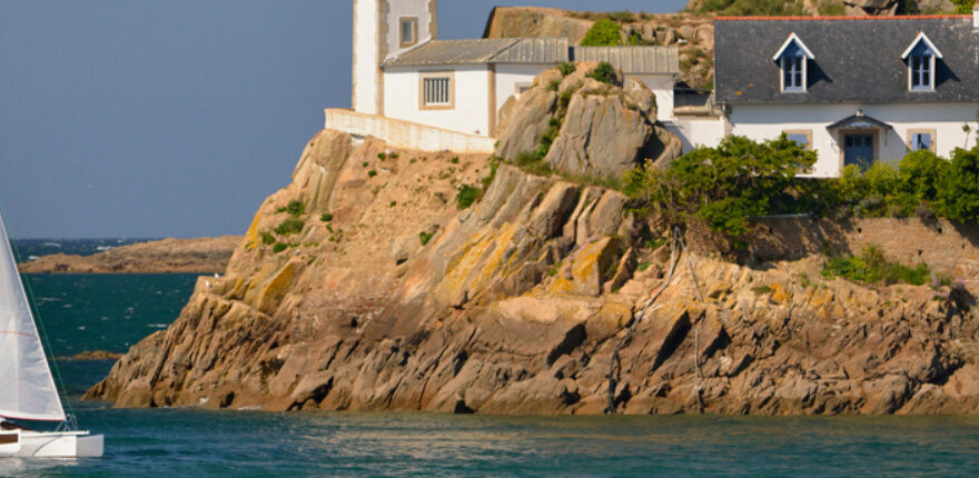 voilier approchant la côte bretonne avec un phare et une petite maison aux volets bleus