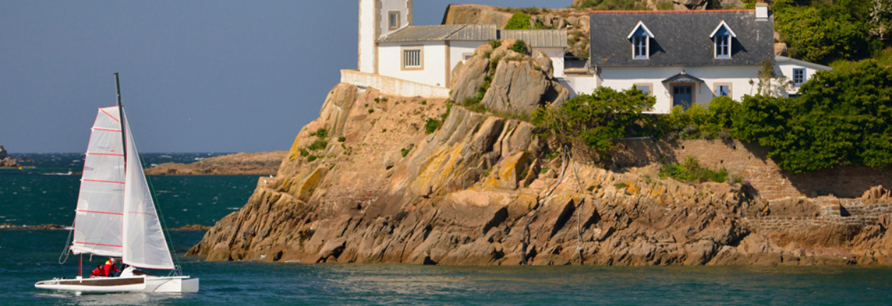 voilier approchant la côte bretonne avec un phare et une petite maison aux volets bleus